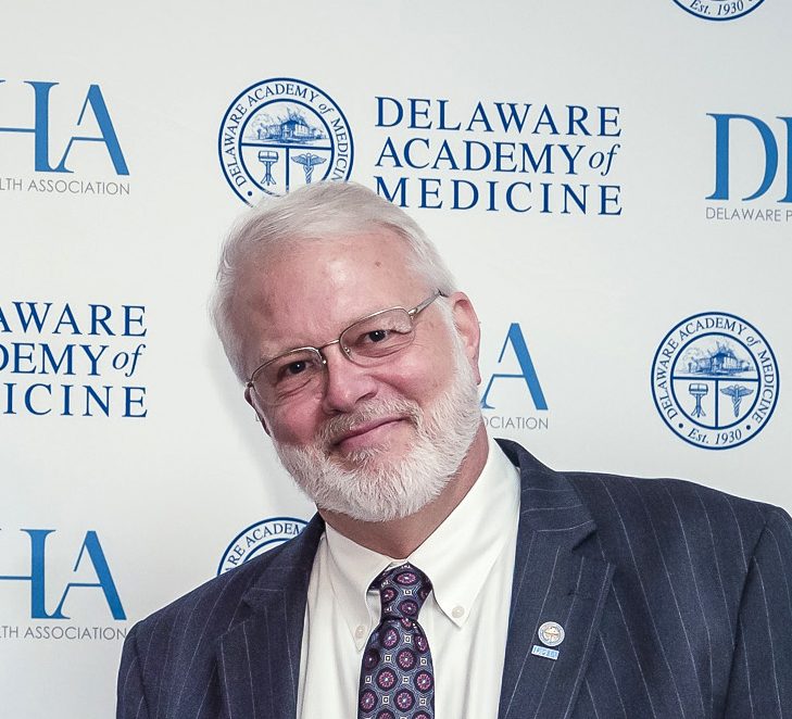 Head and shoulders of a smiling man with glasses and a white beard, wearing a suit and tie, standing in front of a Delaware Academy of Medicine - Delaware Public Health Association step-and-repeat background